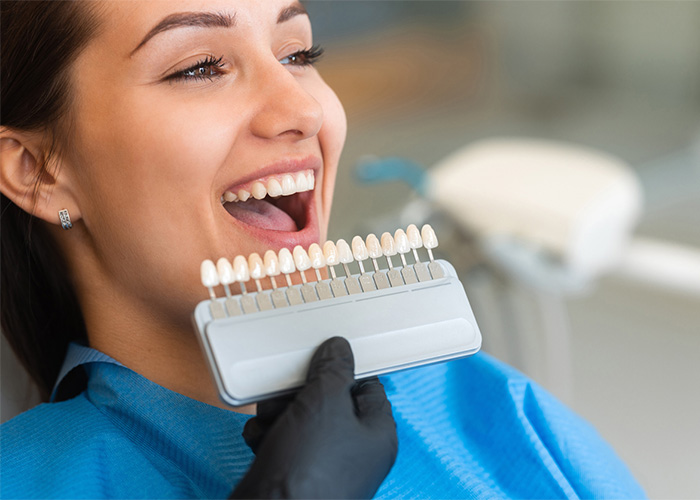 A woman in a dental chair smiles widely as a dentist holds up a set of tooth shade guides next to her mouth. She wears a blue dental bib, and the dentist wears black gloves.